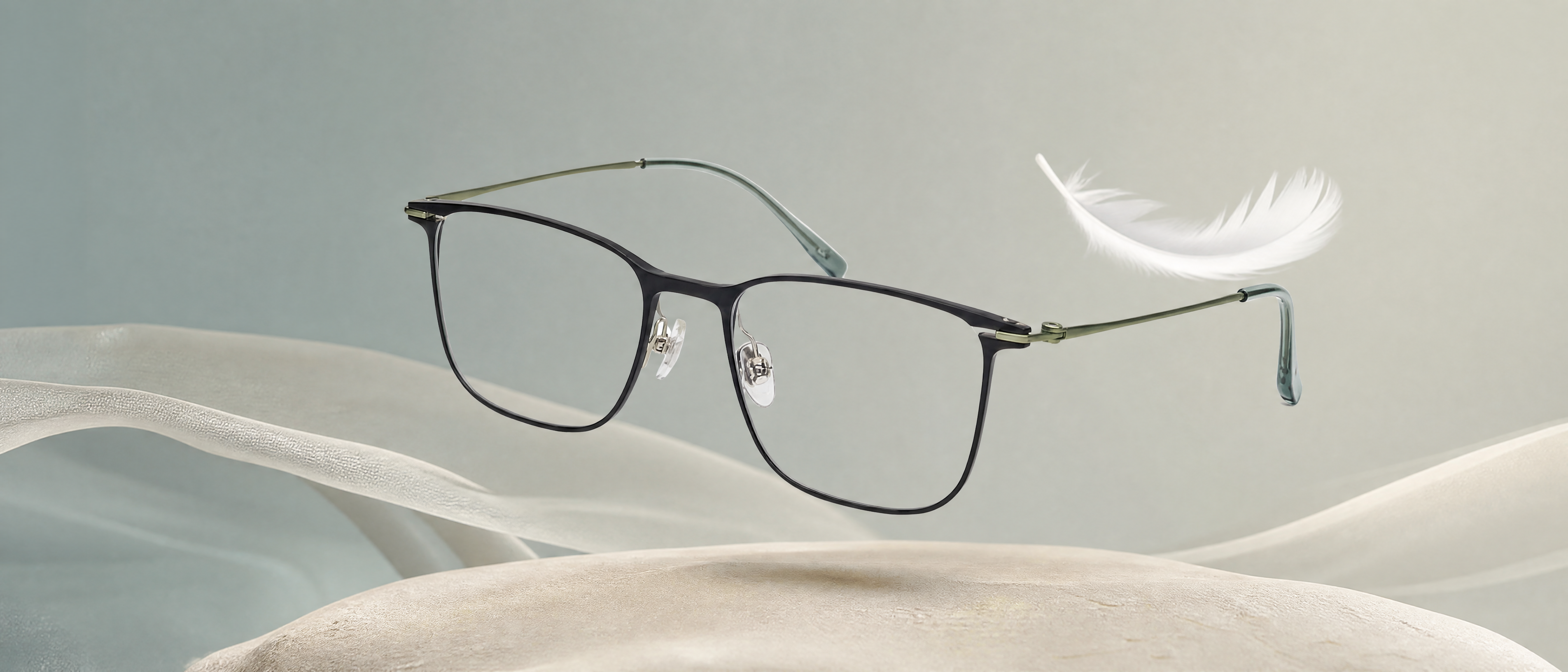 A pair of slim metal eyeglasses with a black front frame and silver temples, floating above draped beige fabric with a white feather graphic on a soft light gray background.