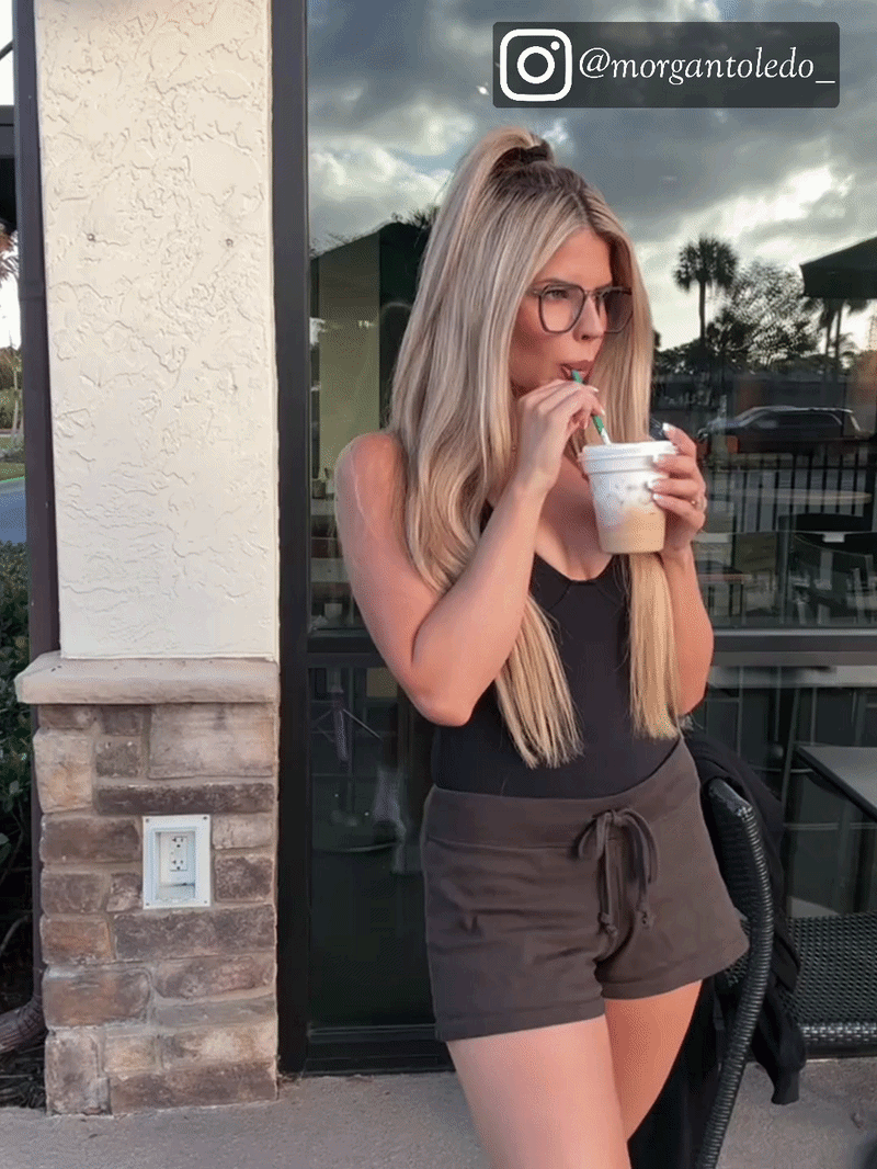 Blonde woman wearing black square eyeglasses drinking iced coffee outdoors, tropical palm tree background, lifestyle product shot
