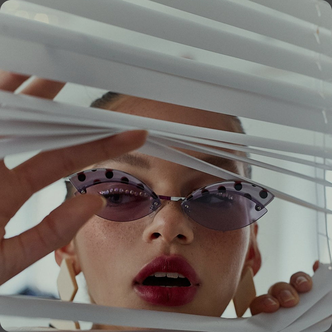 Person wearing purple sunglasses looking through white blinds